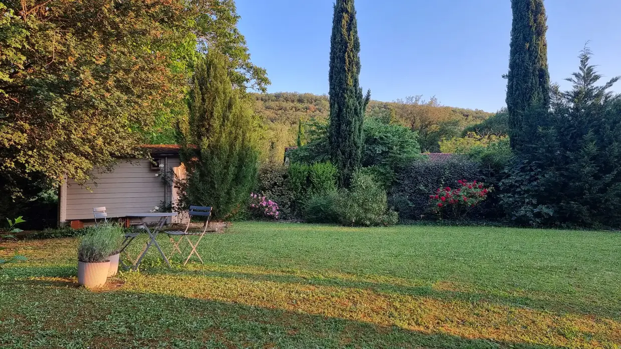 l'heure douce, on voit le jardin depuis la fenêtre du bureau avec le causse à l'arrière plan, les cyprès, les rosiers, sur la gauche une cabane, une table dressée à côté de deux pots de lavande sur la pelouse