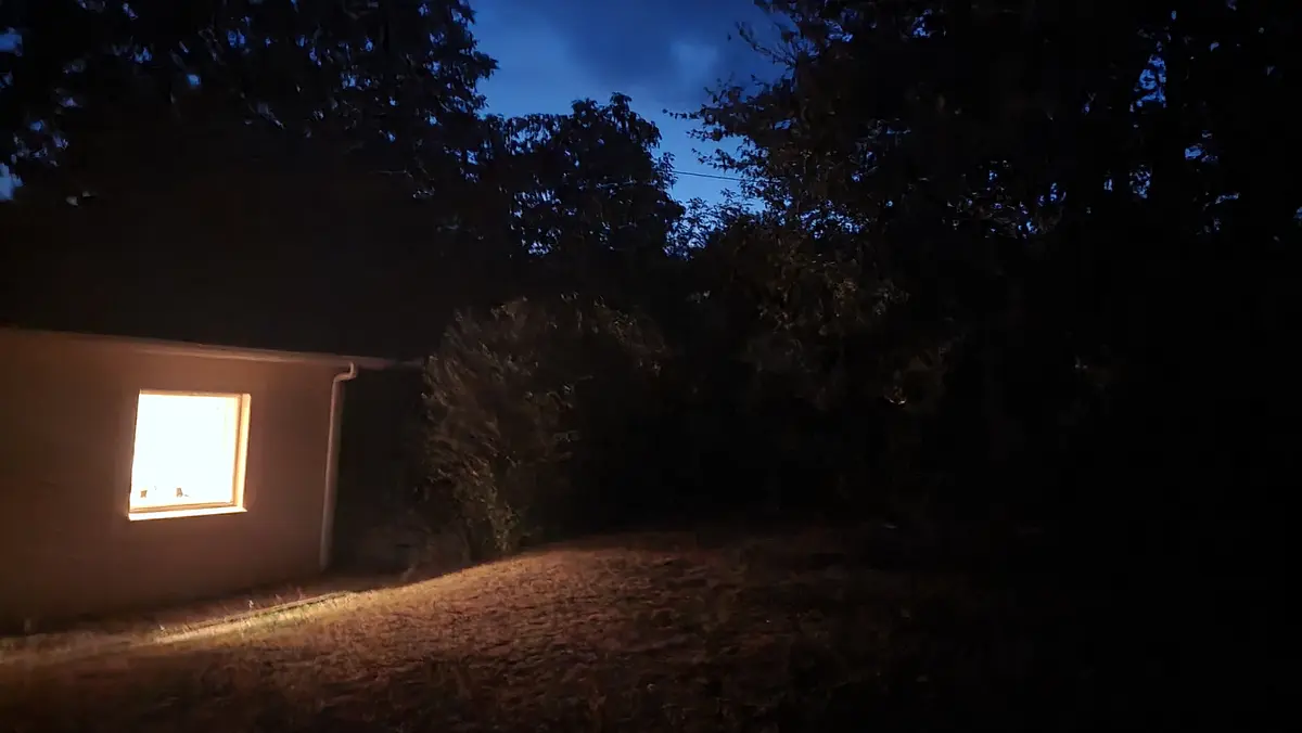 on voit la petite fenêtre du bureau éclairée à l'aube, on devine à l'arrière plan les arbres du jardin qui se découpent sur le ciel et devant un talus à l'herbe sèche